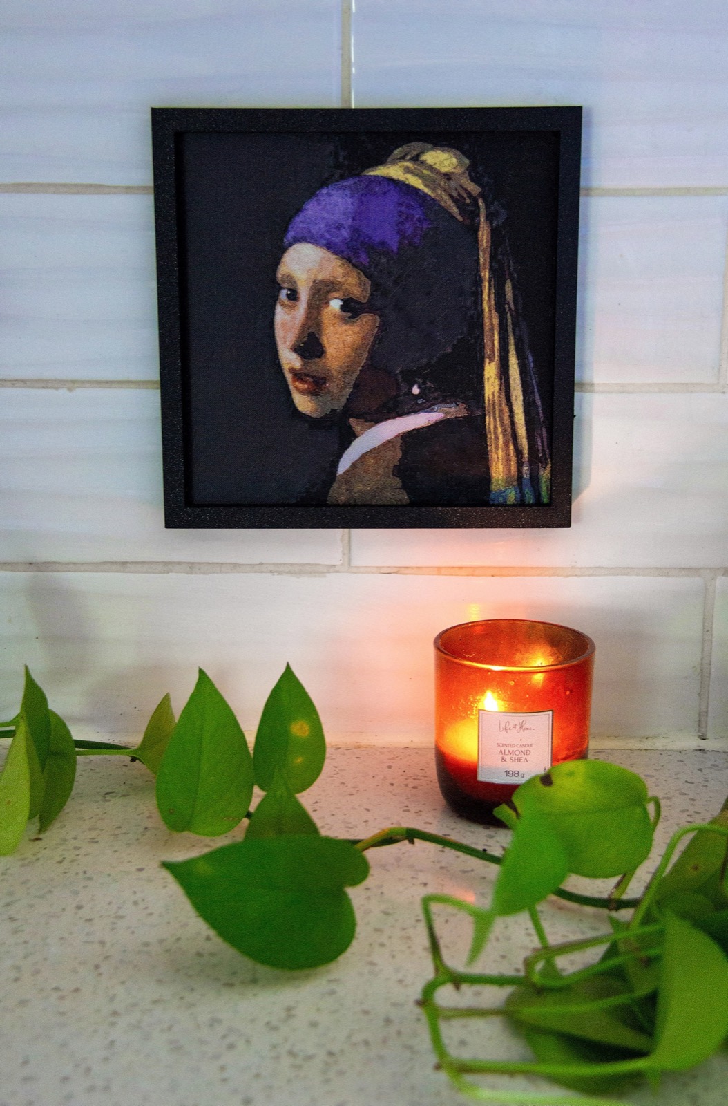 A framed Girl with a Pearl Earring print on a tiled wall above a lit candle